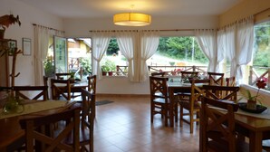 A dining area with wooden tables and chairs, a large window with curtains, and a hanging light fixture. - Tierra Sureña (San Carlos de Bariloche)