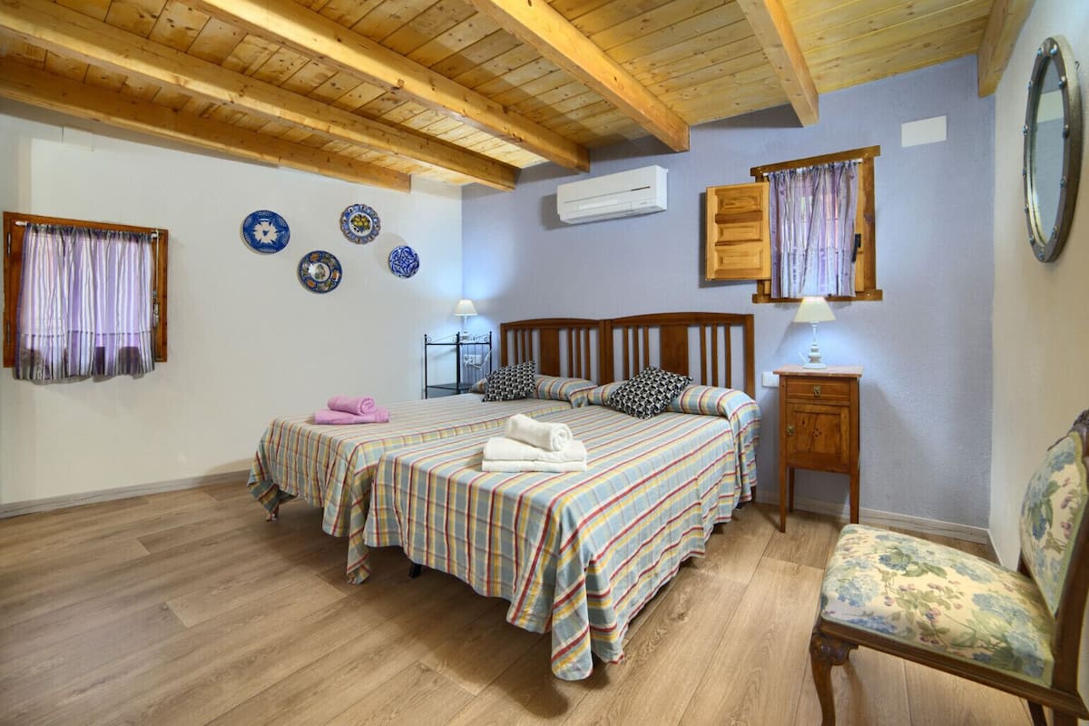 Ein Schlafzimmer mit zwei Betten, Holzbalken an der Decke, einem kleinen Nachttisch, einem Sessel und dekorativen Tellern an der Wand.
