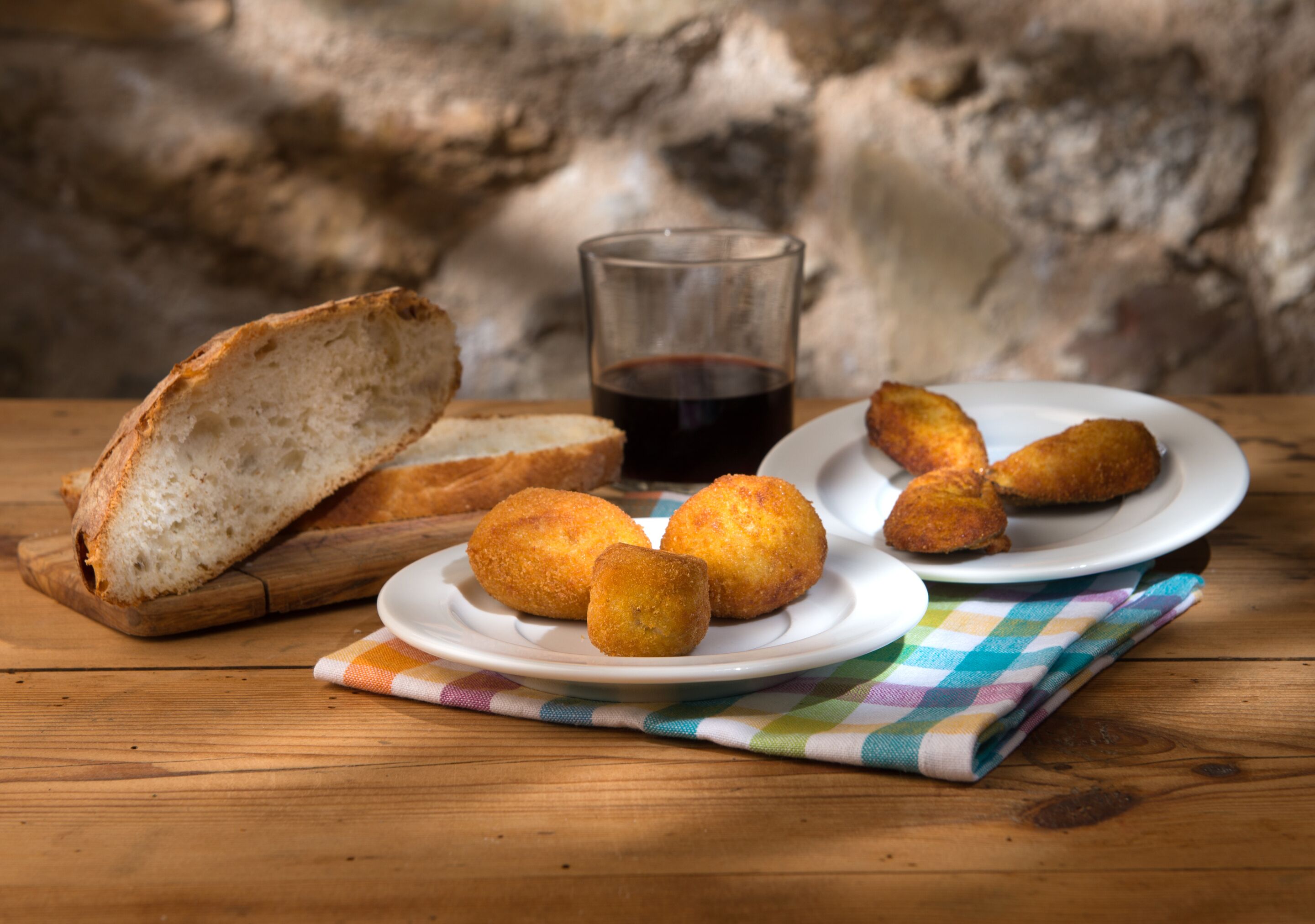 Una mesa de madera con un mantel a cuadros, una copa de vino tinto y platos de croquetas y pan.