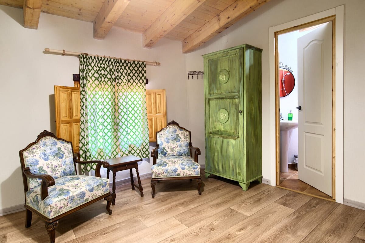 Ein Zimmer mit Holzboden, zwei gepolsterte Sessel mit Blumenmuster sowie ein grünes Schranksystem und eine Tür zu einem Badezimmer.