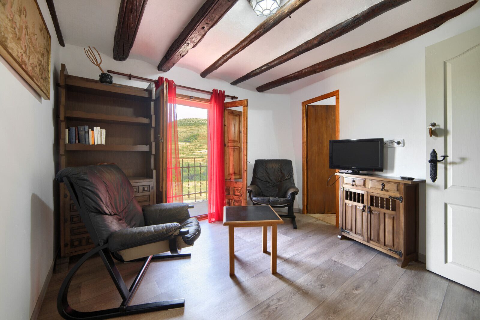 A cozy living room with a wooden floor, a leather armchair, a wooden table, a television on a wooden cabinet, and a bookshelf.