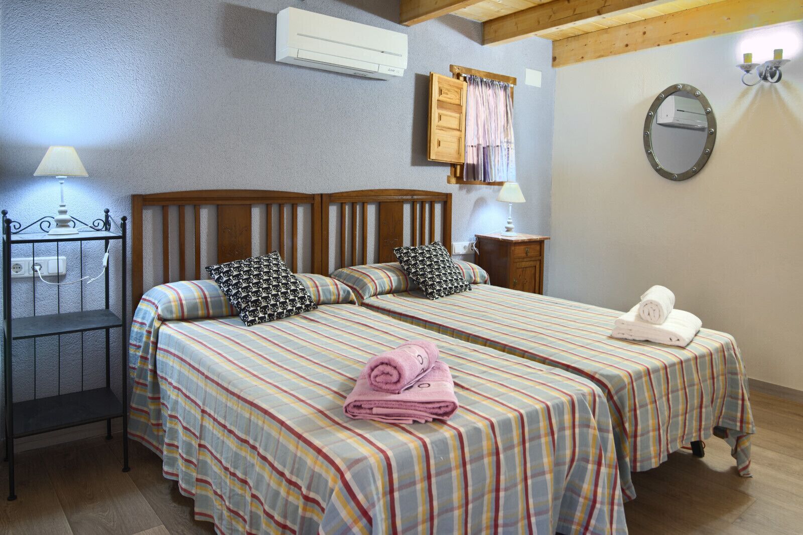 Habitación doble con dos camas, cada una con ropa de cama y almohadas a rayas, cabecero de madera, mesita de noche pequeña con lámpara y espejo montado en la pared.