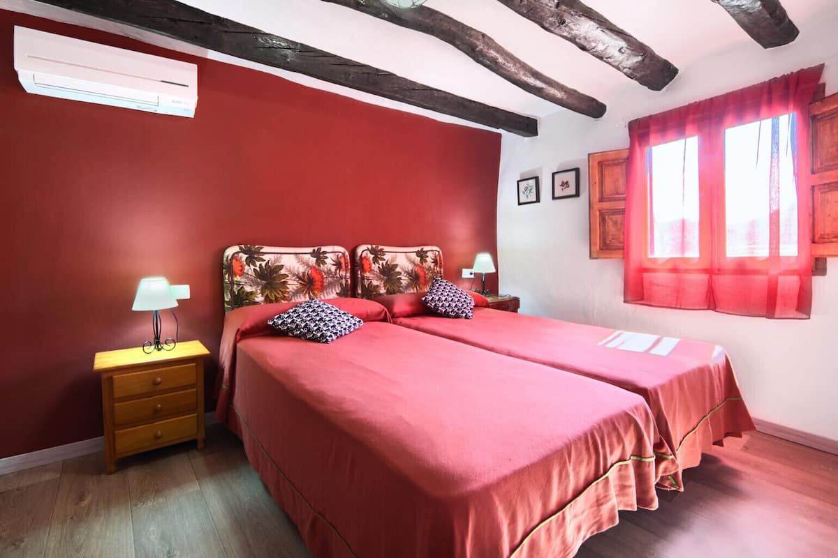Ein Zimmer mit zwei Betten, hölzernen Nachttischen, einer roten Wand, einem Fenster mit roten Vorhängen und einer Decke mit sichtbaren Holzbalken.