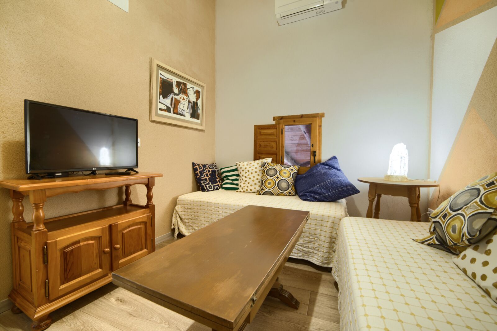 Une petite pièce agréable, avec un meuble TV en bois, un lit recouvert d’une literie à motifs, une petite table et un climatiseur fixé au mur.