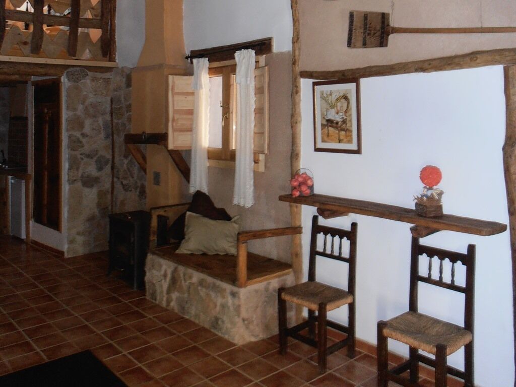 Un interior rústico con un banco de piedra, sillas de madera, una estantería de madera con un jarrón y un cuadro en la pared.