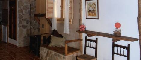Un interior rústico con un banco de piedra, sillas de madera, una estantería de madera con un jarrón y un cuadro en la pared.