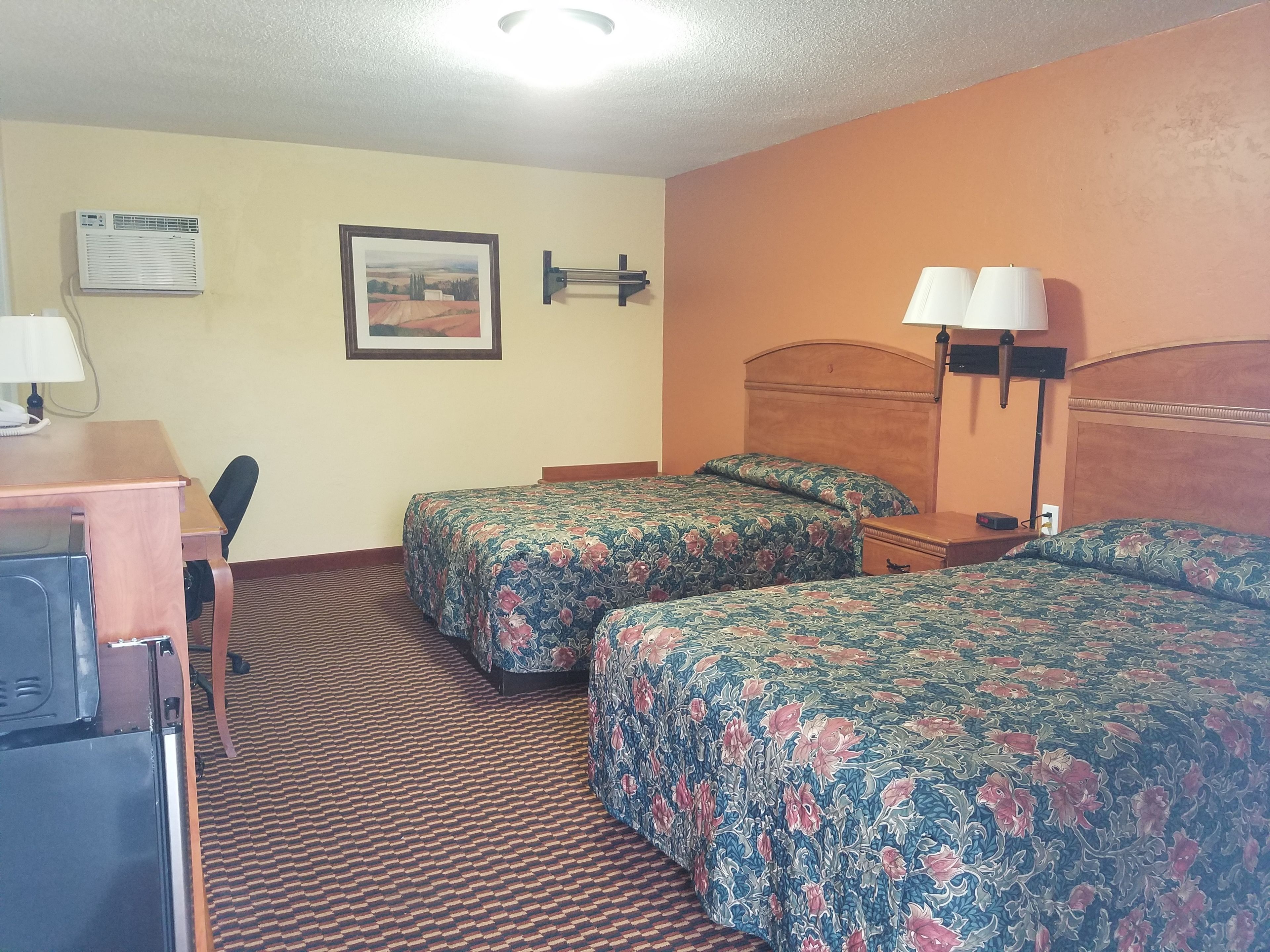 Habitación de hotel con dos camas, un escritorio, una silla, una lámpara y un cuadro en la pared.