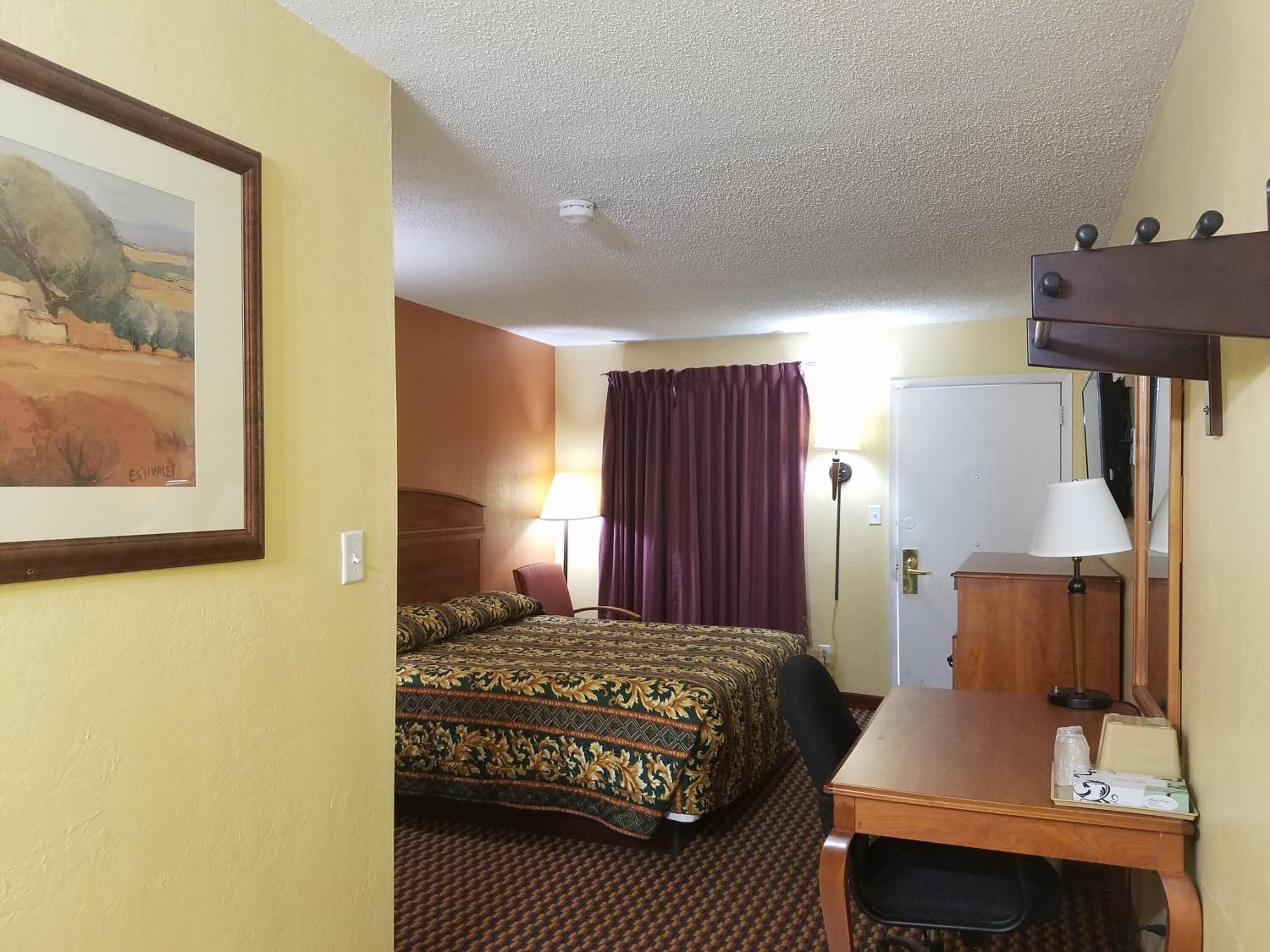 A hotel room with a bed, a desk, a chair, a lamp, a wardrobe, and a painting on the wall.