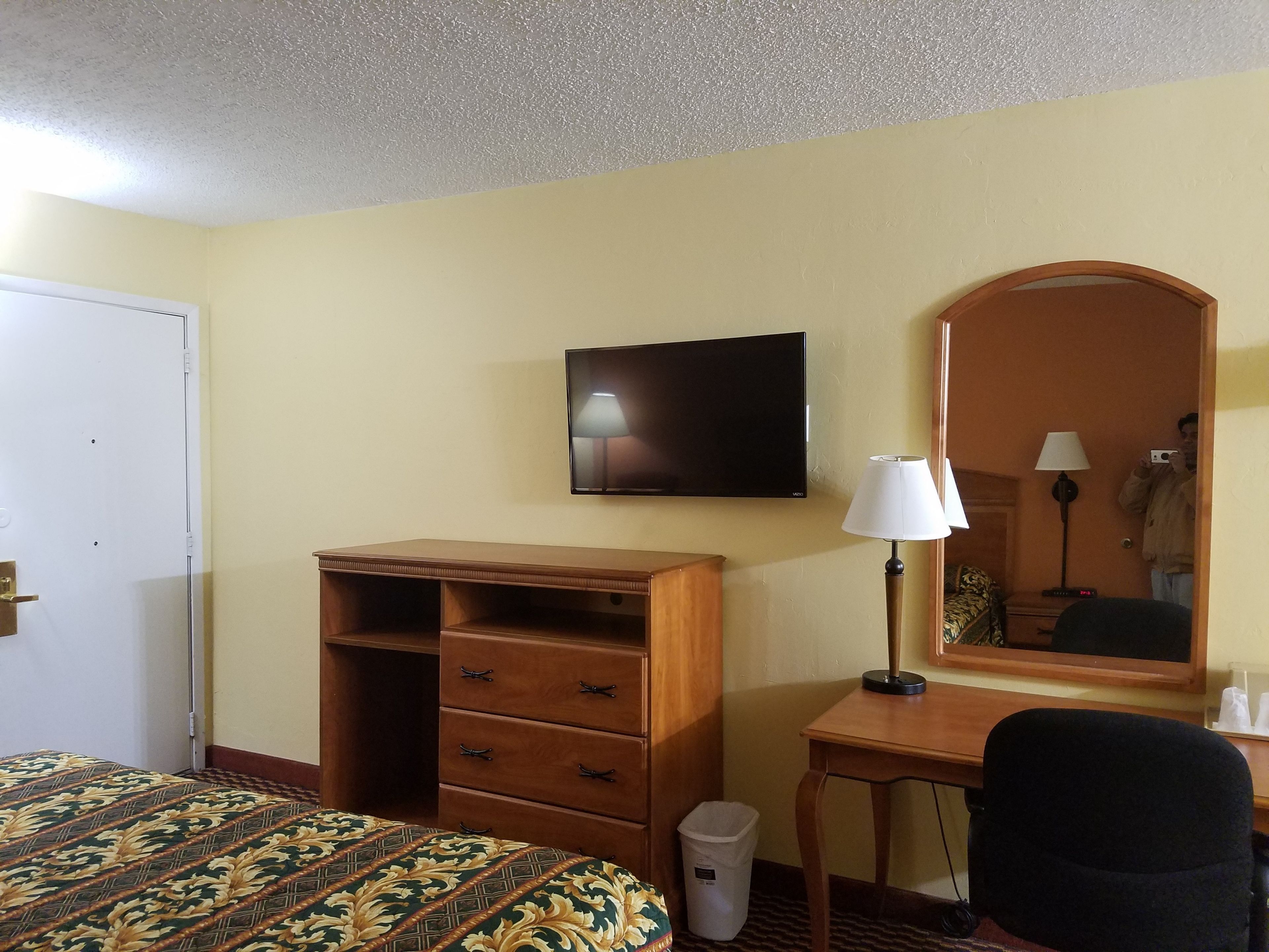 A hotel room with a bed, a wooden dresser, a desk with a lamp, a television, and a mirror.