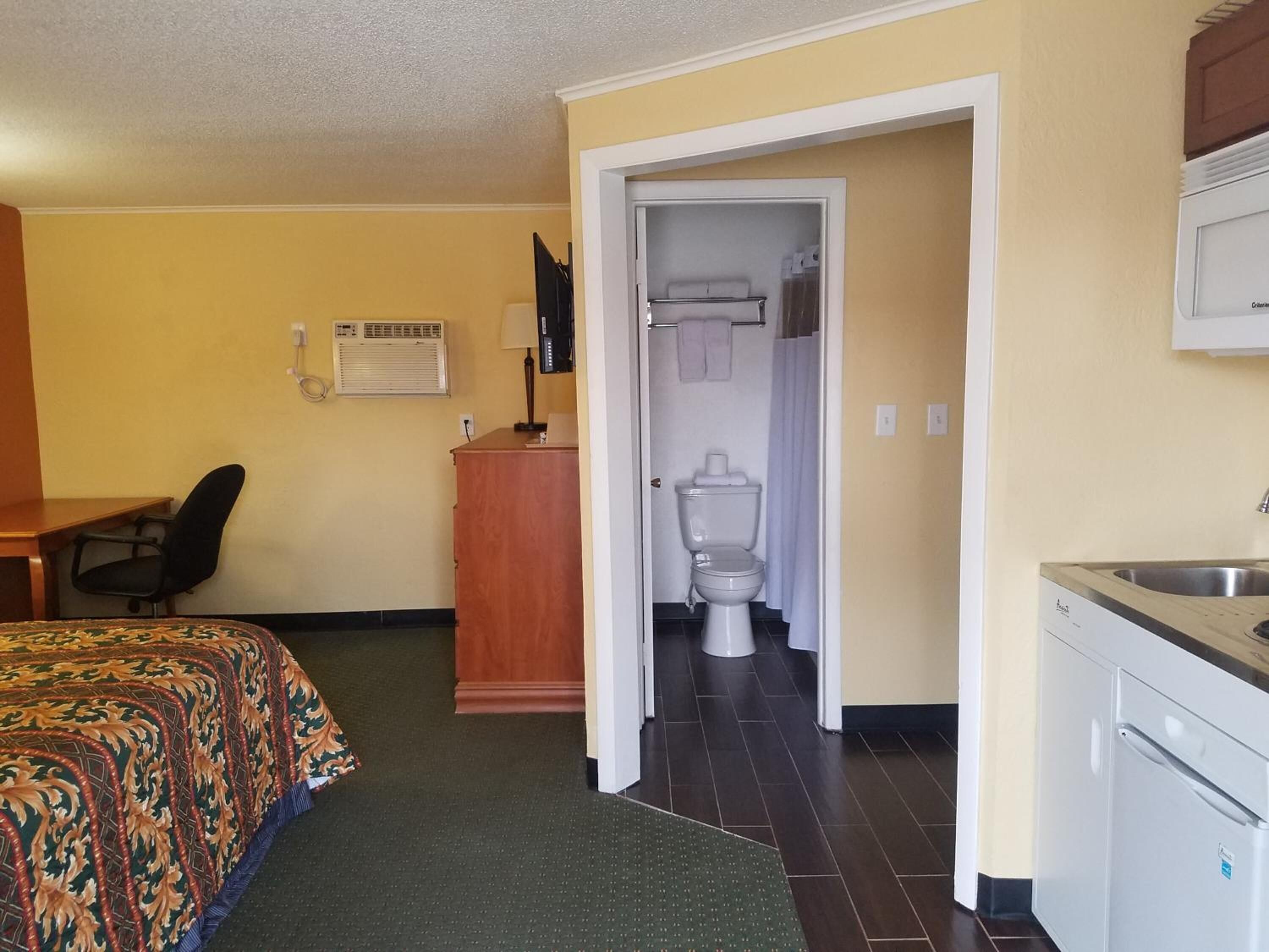 A hotel room with a bed, desk, kitchenette, and a bathroom visible through an open door.
