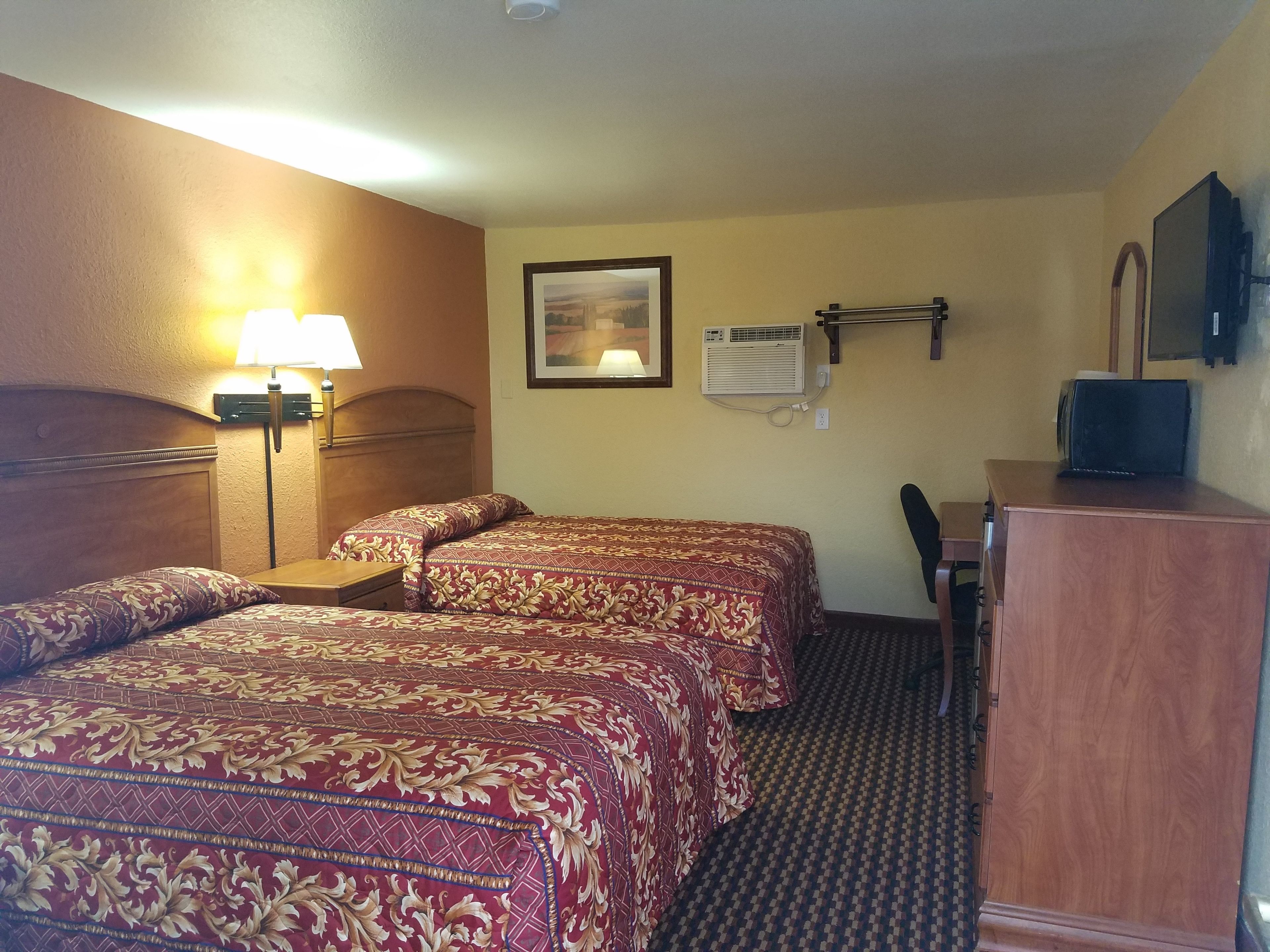 A hotel room with two beds, a desk, a chair, a lamp, an air conditioning unit, and a television.