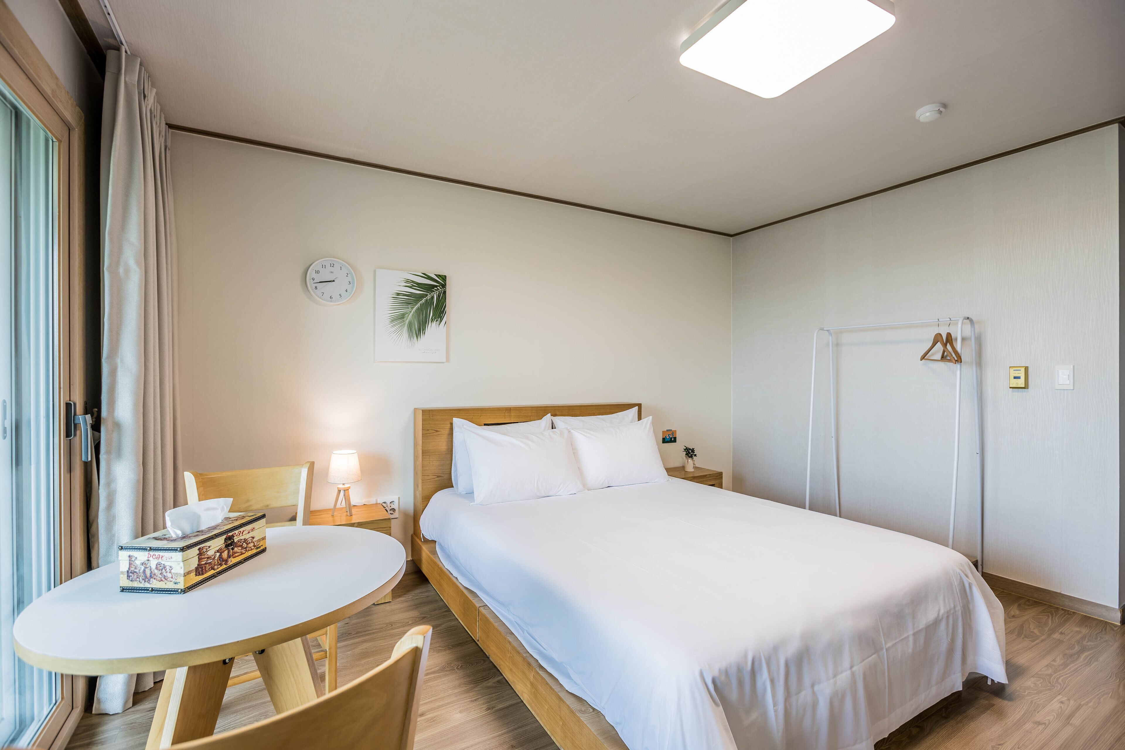 A hotel room with a bed, a table, and a chair. There is a clock and a plant on the wall.