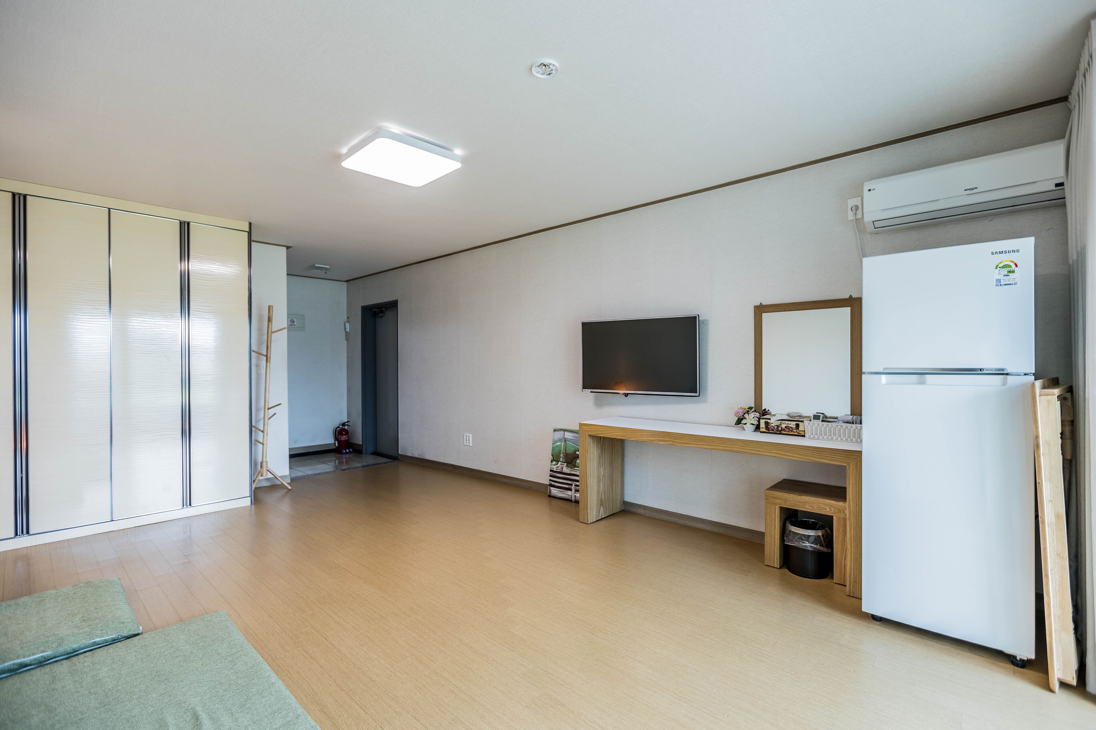 A modern apartment with a wooden floor, a flat-screen TV mounted on the wall, a white refrigerator, and a wooden desk with a small trash bin underneath.