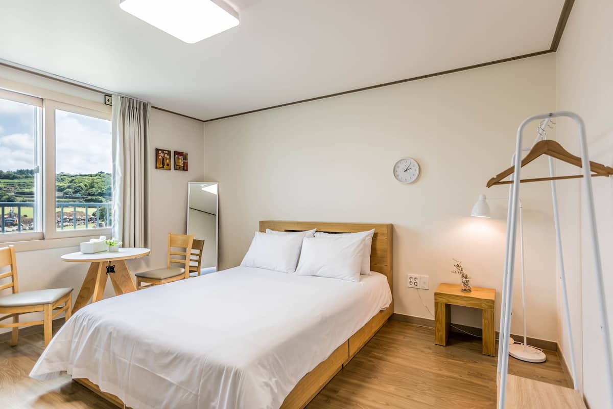 Habitación de hotel con cama, una mesita con sillas, una mesita de noche, una lámpara colgante y vistas al exterior.
