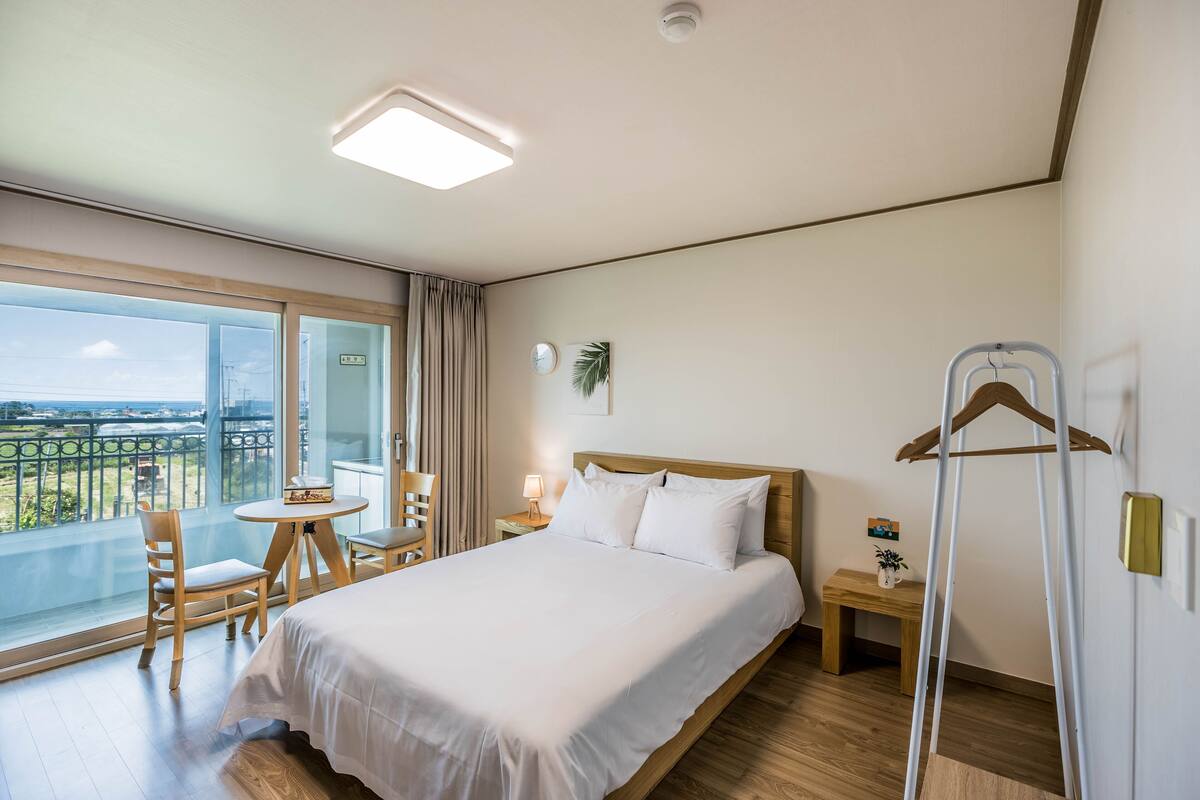 Habitación de hotel con una cama grande, un balcón con vistas, una mesita con una silla y una mesita de noche con una lámpara.