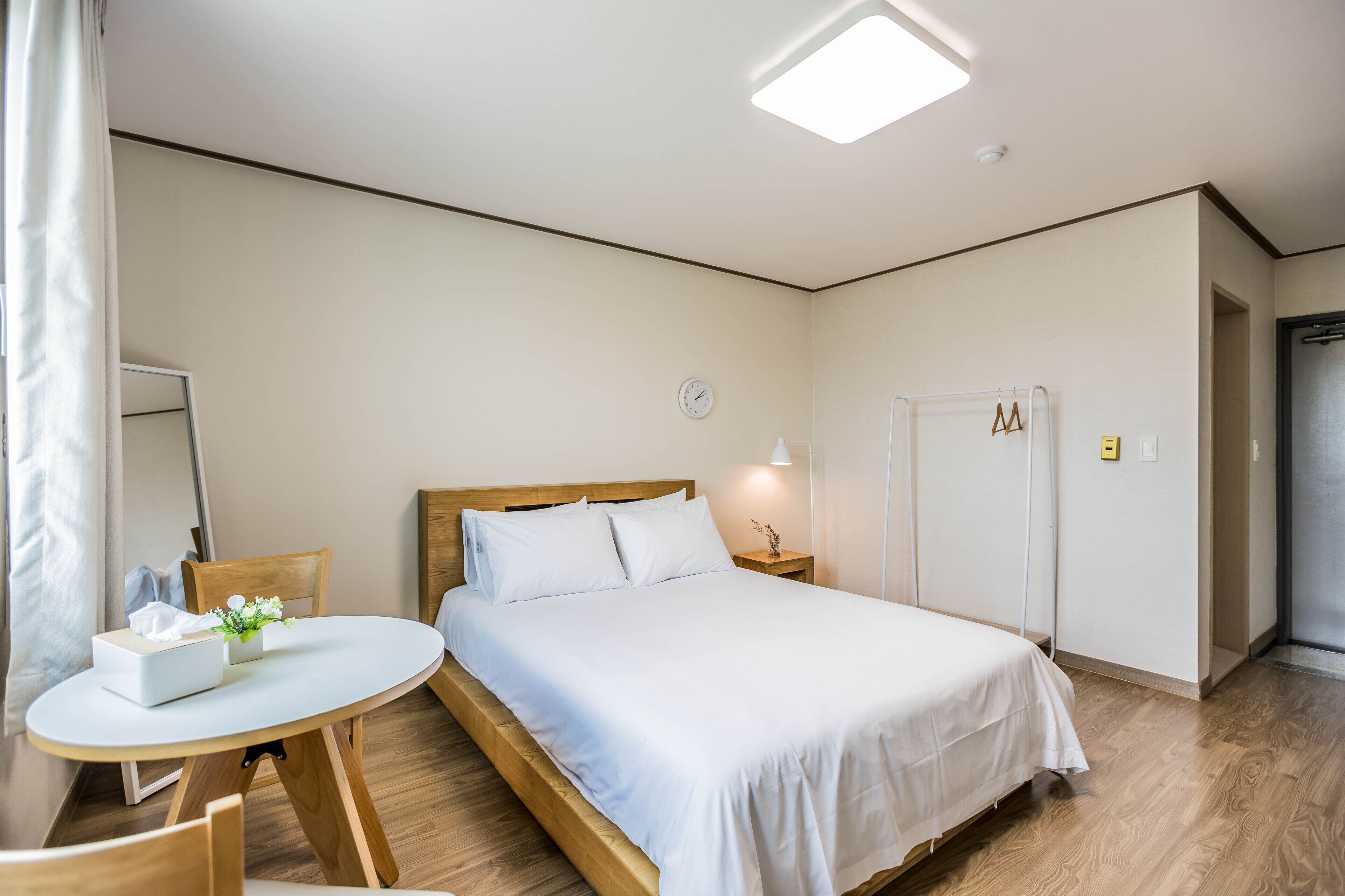A bedroom with a wooden bed, white bedding, a bedside table with a lamp, a small table with a box and flowers, and a mirror on the wall.