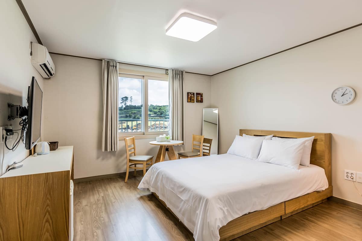 Habitación de hotel con cama, escritorio, silla, mesita, ventana con cortinas y un reloj en la pared.