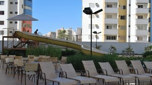 Zona de piscina con sillas de descanso y un tobogán, rodeada de edificios.