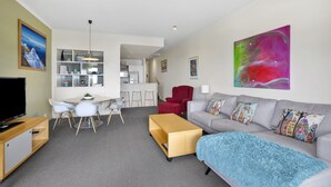 A modern living room with a grey sofa, a wooden coffee table, and a TV on a stand. There is a dining area with a white table and chairs, and a kitchen in the background. - Lorne Beachfront Accommodation (Lorne)