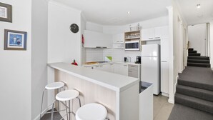A modern kitchen with white cabinetry, a central island with bar stools, a microwave, and a refrigerator. - Lorne Beachfront Accommodation (Lorne)