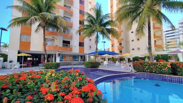 A hotel with a swimming pool, outdoor seating, and palm trees. -  Apartamento Jardins das Thermas - Achei Férias (com roupa de cama) (Caldas Novas)
