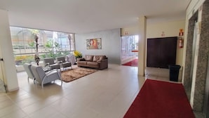 A spacious living area with a red carpet, a brown leather sofa, grey armchairs, and a large window with plants. -  Apartamento Jardins das Thermas - Achei Férias (com roupa de cama) (Caldas Novas)