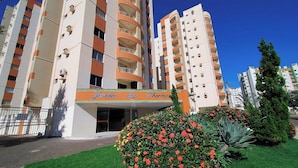 A multi-story building with balconies, a sign reading "Jardins da Terra," and a well-maintained garden with red flowers. -  Apartamento Jardins das Thermas - Achei Férias (com roupa de cama) (Caldas Novas)