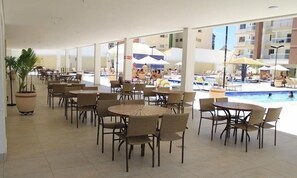 A spacious indoor area with multiple tables and chairs, a pool area with umbrellas, and a view of a building in the background.