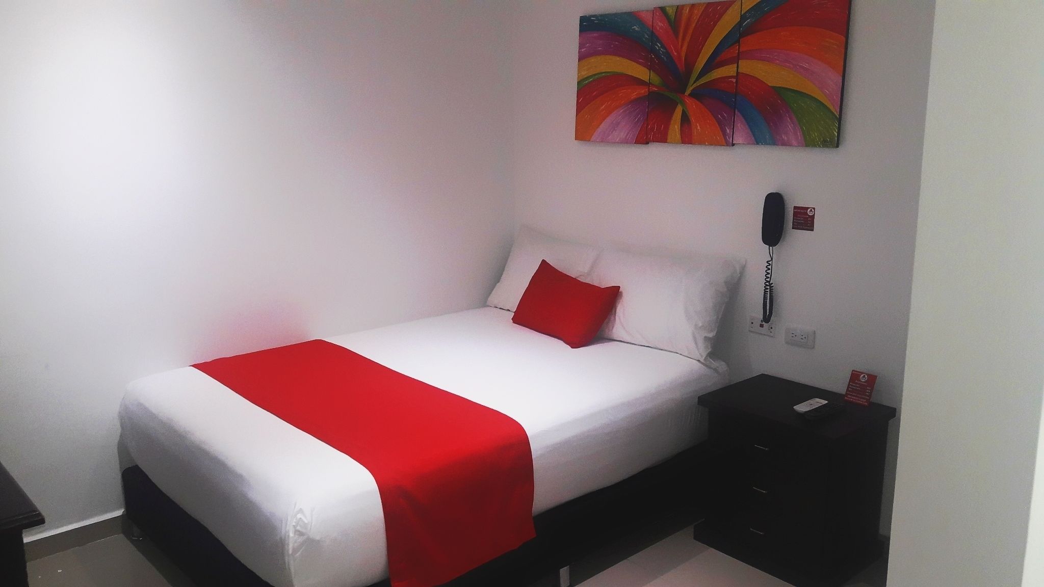 A single bed with a white and red bedspread, a red pillow, and a black nightstand with a phone.