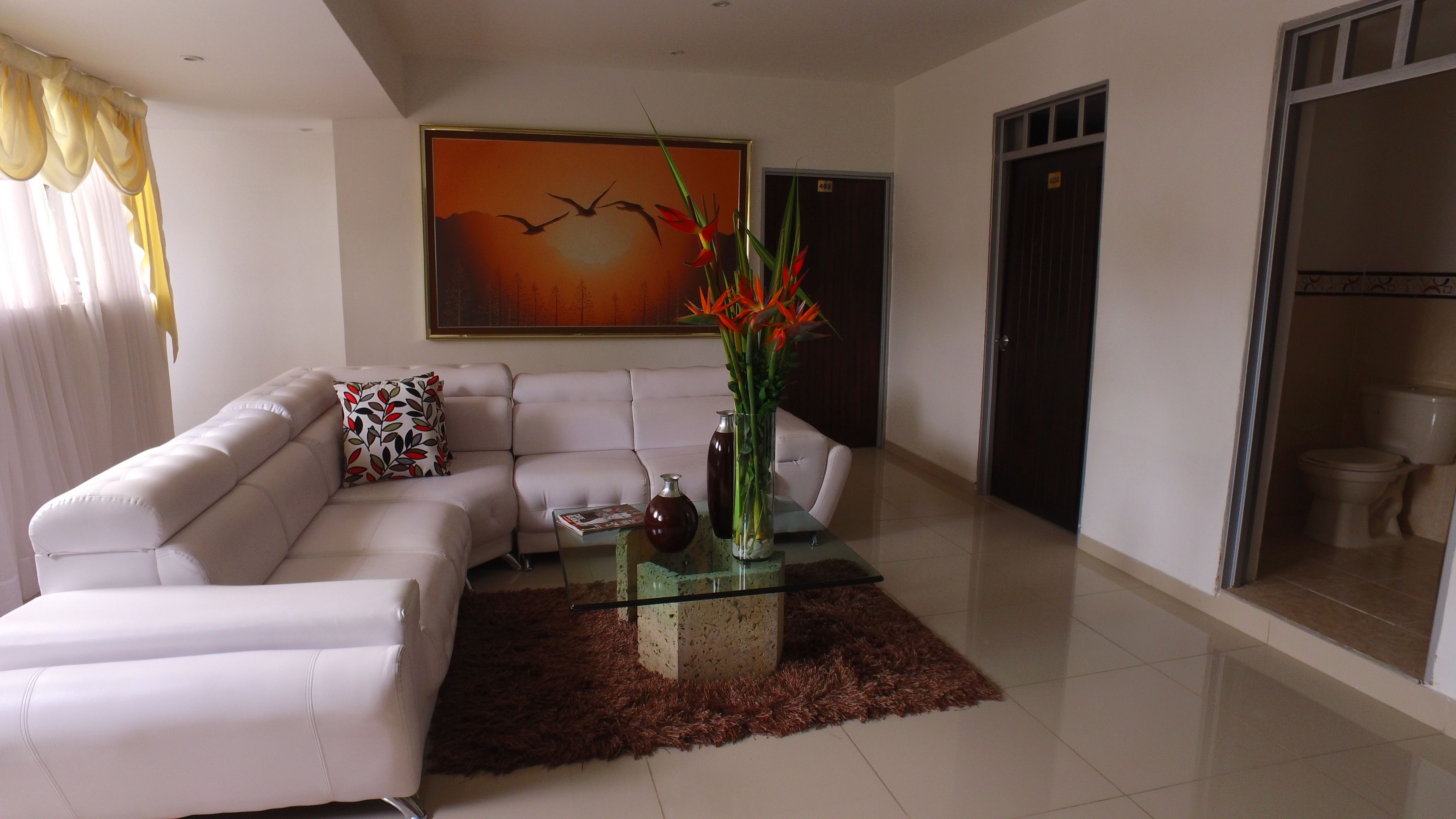 A modern living room with a white leather sofa, a glass coffee table with decorative items, a large painting on the wall, and a view of the adjacent bathroom.