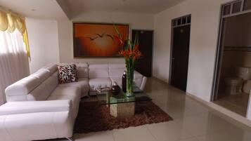 A modern living room with a white leather sofa, a glass coffee table with decorative items, a large painting on the wall, and a view of the adjacent bathroom.