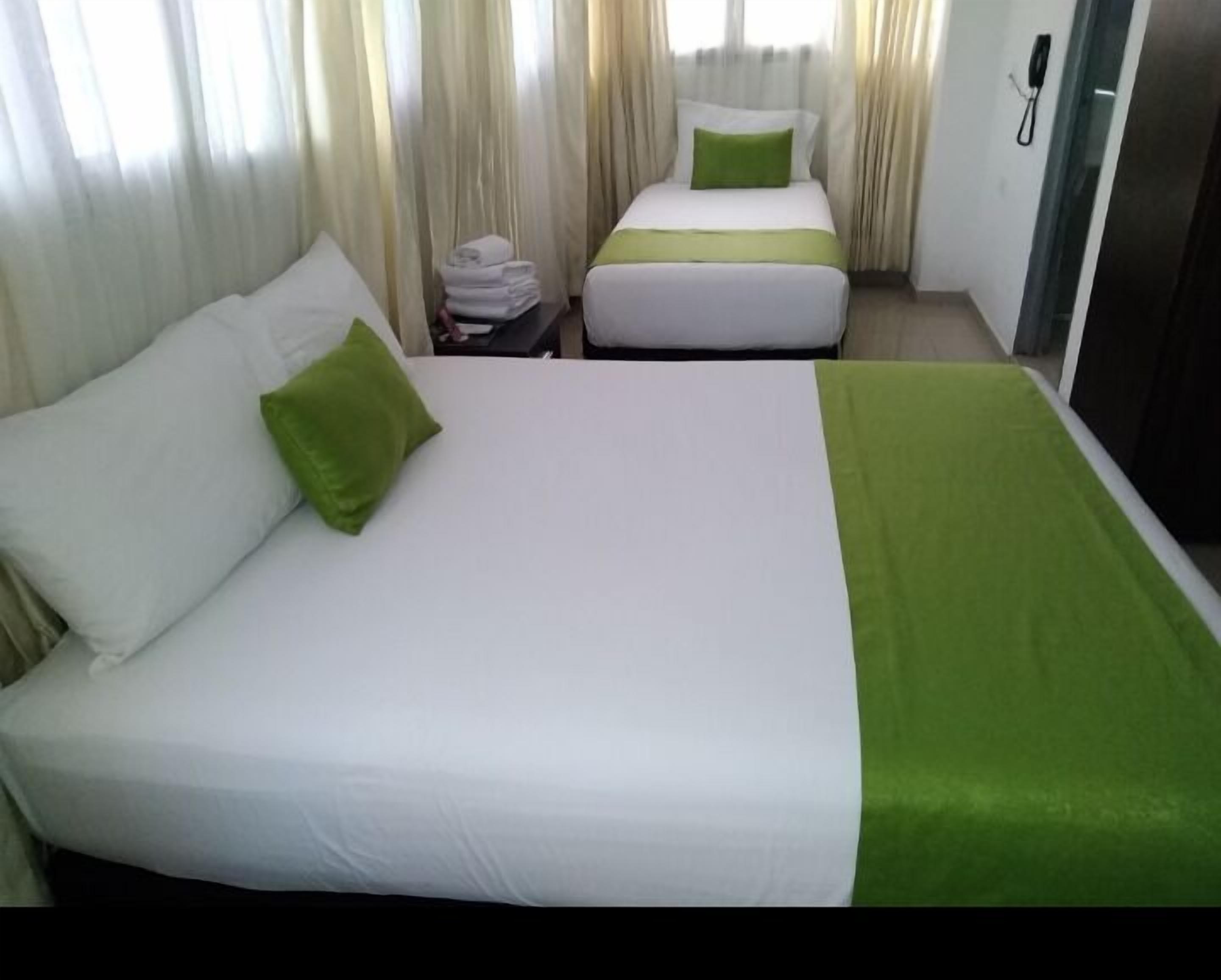 A hotel room with two beds, each with white bedding and green accents. There is a small table with a stack of towels and a telephone on the wall.