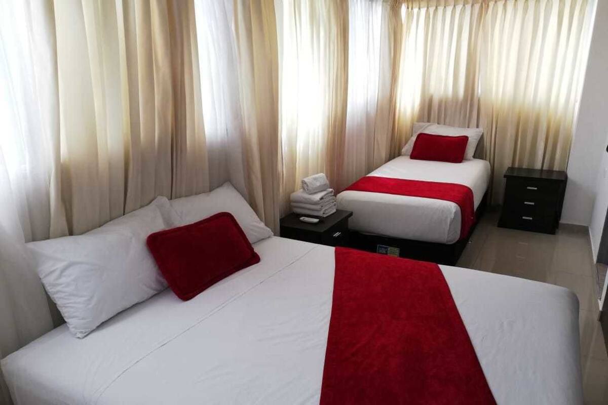 A hotel room with two beds, each with white bedding and red accents, a small bedside table with a remote control, and a dark-colored nightstand.