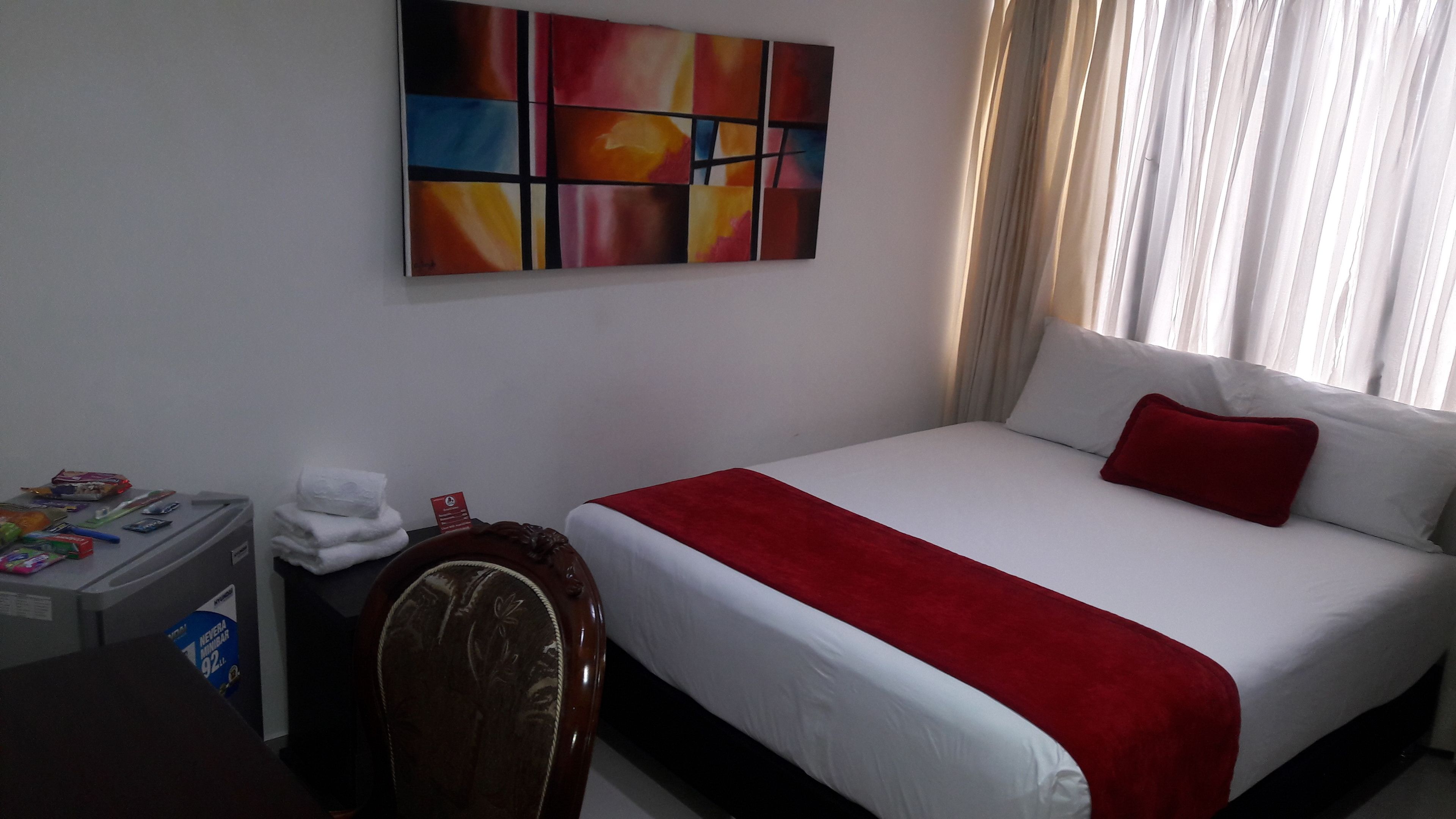 A hotel room with a large bed, a red throw blanket, a white pillow, a small table with toiletries, and a colorful abstract painting on the wall.