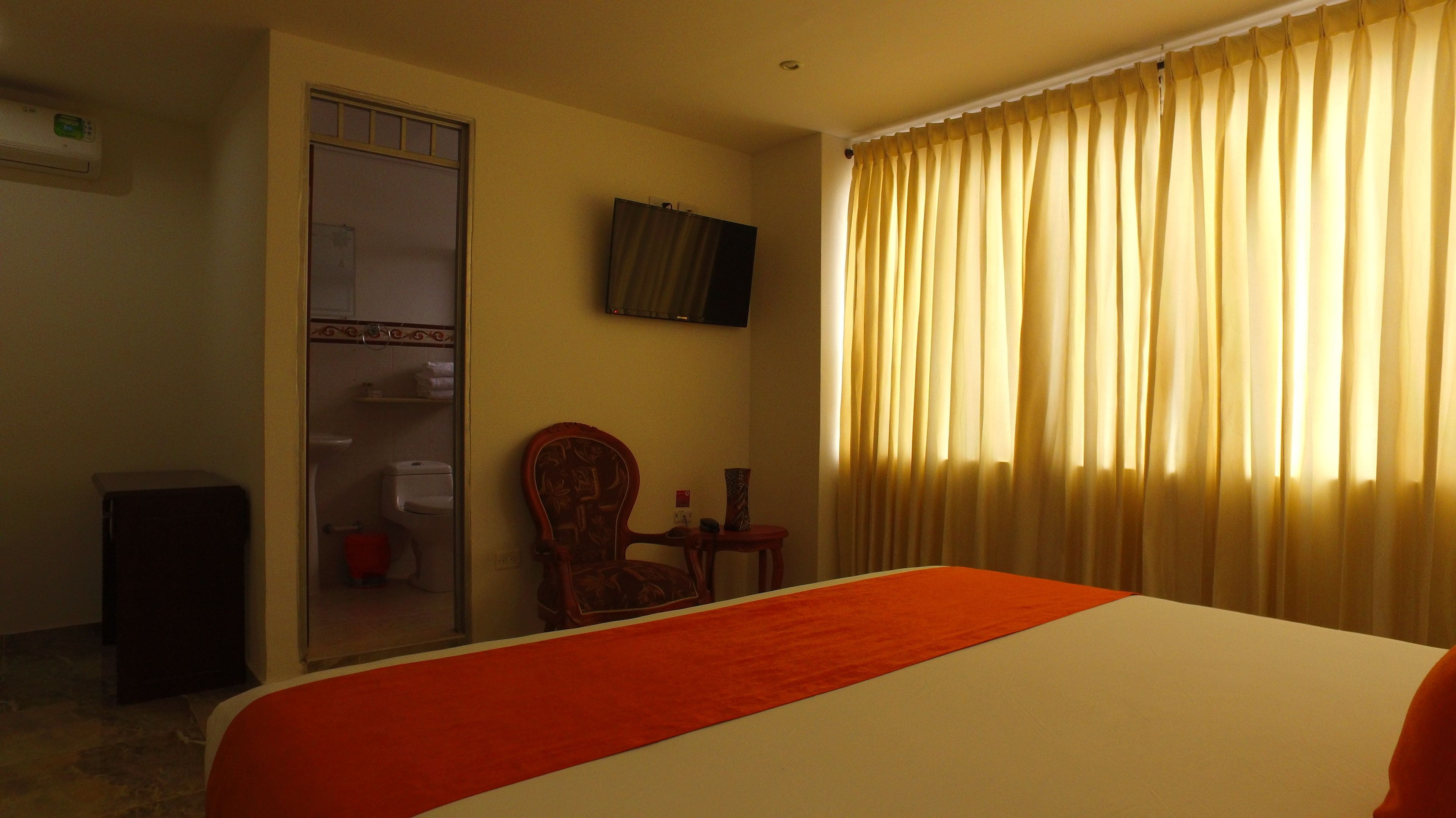 Une chambre d’hôtel avec un lit, une chaise, une petite table, un téléviseur et une fenêtre avec des rideaux.
