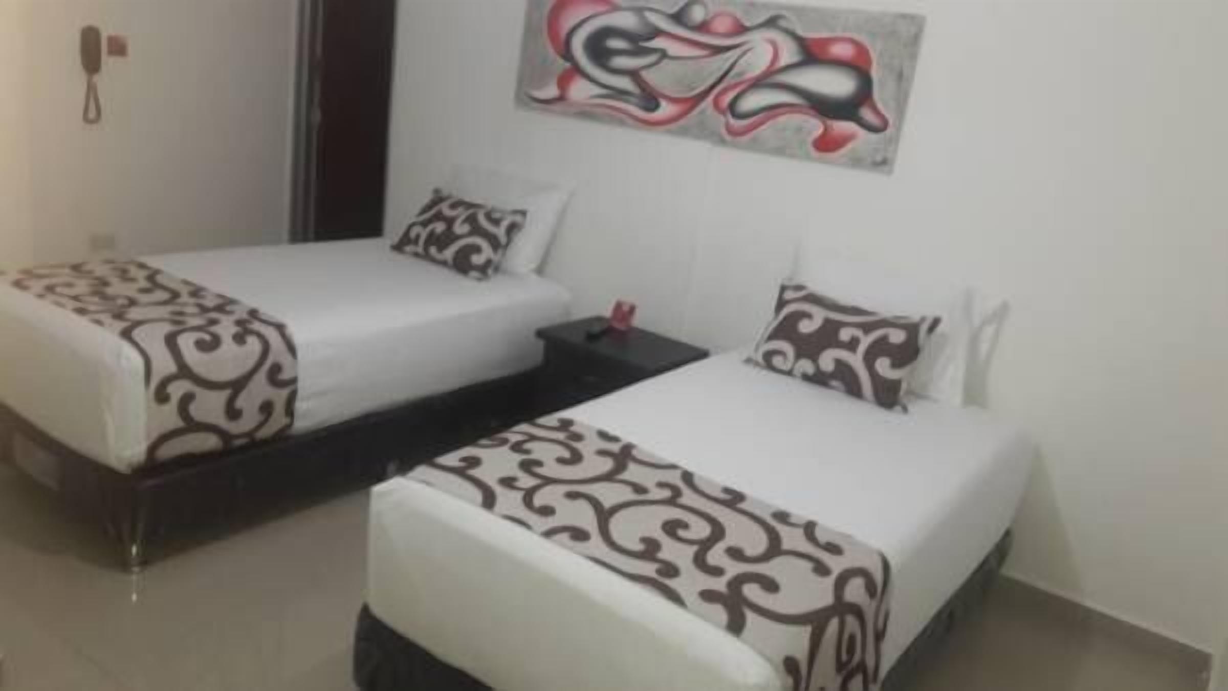 Habitación de hotel con dos camas individuales, cada una con ropa de cama y almohadas estampadas. Hay una mesita de noche pequeña con una lámpara y un objeto rojo encima. La habitación tiene una pared blanca lisa con una obra de arte abstracta sobre las camas.