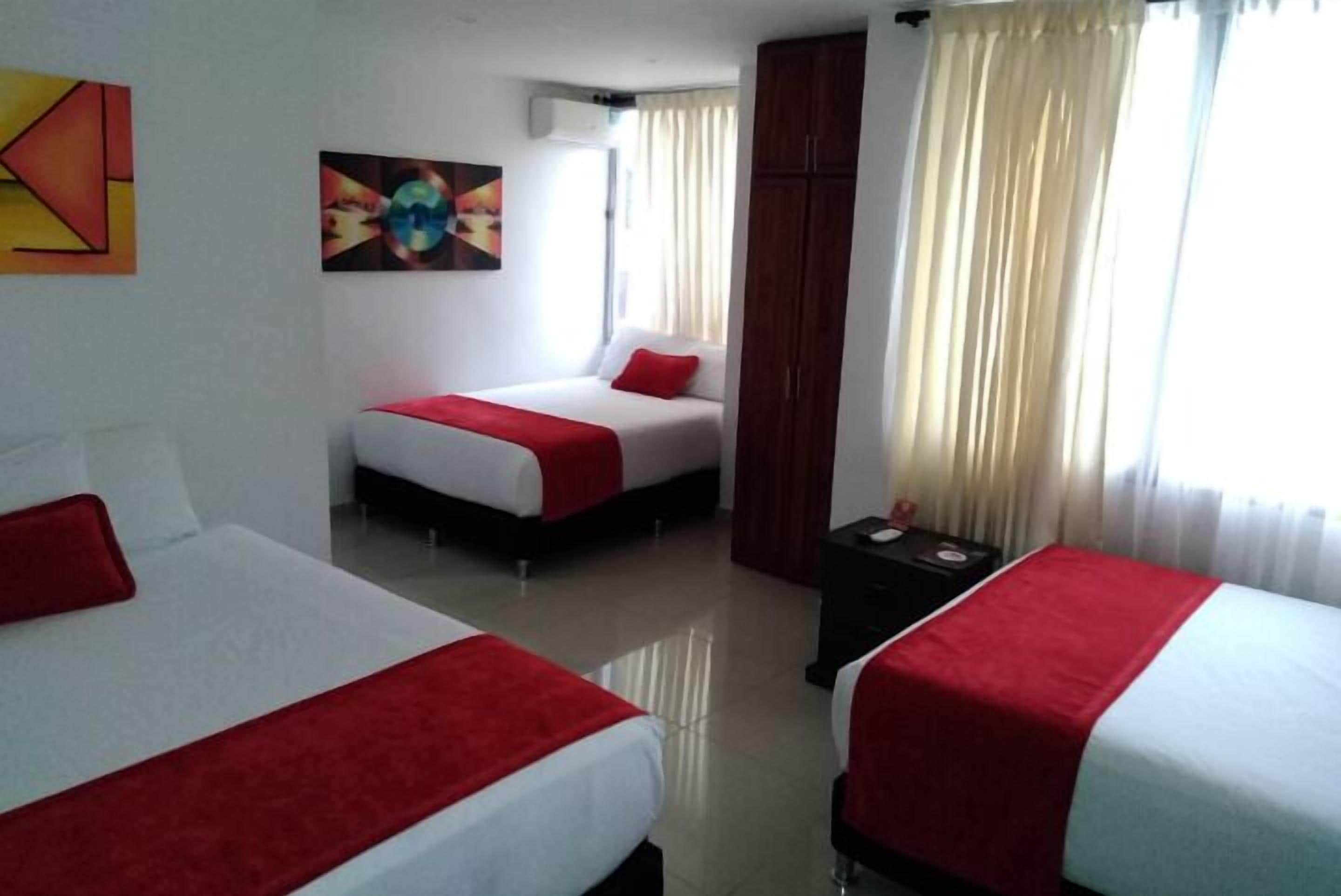 A hotel room with two beds, each with a red blanket and white sheets. There is a painting on the wall, a window with curtains, and a wooden wardrobe.