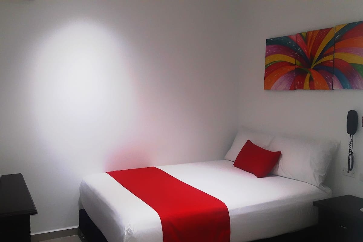 A hotel room with a bed, a red and white bedspread, a white pillow, a red pillow, a black nightstand, a white nightstand, and a colorful abstract painting on the wall.