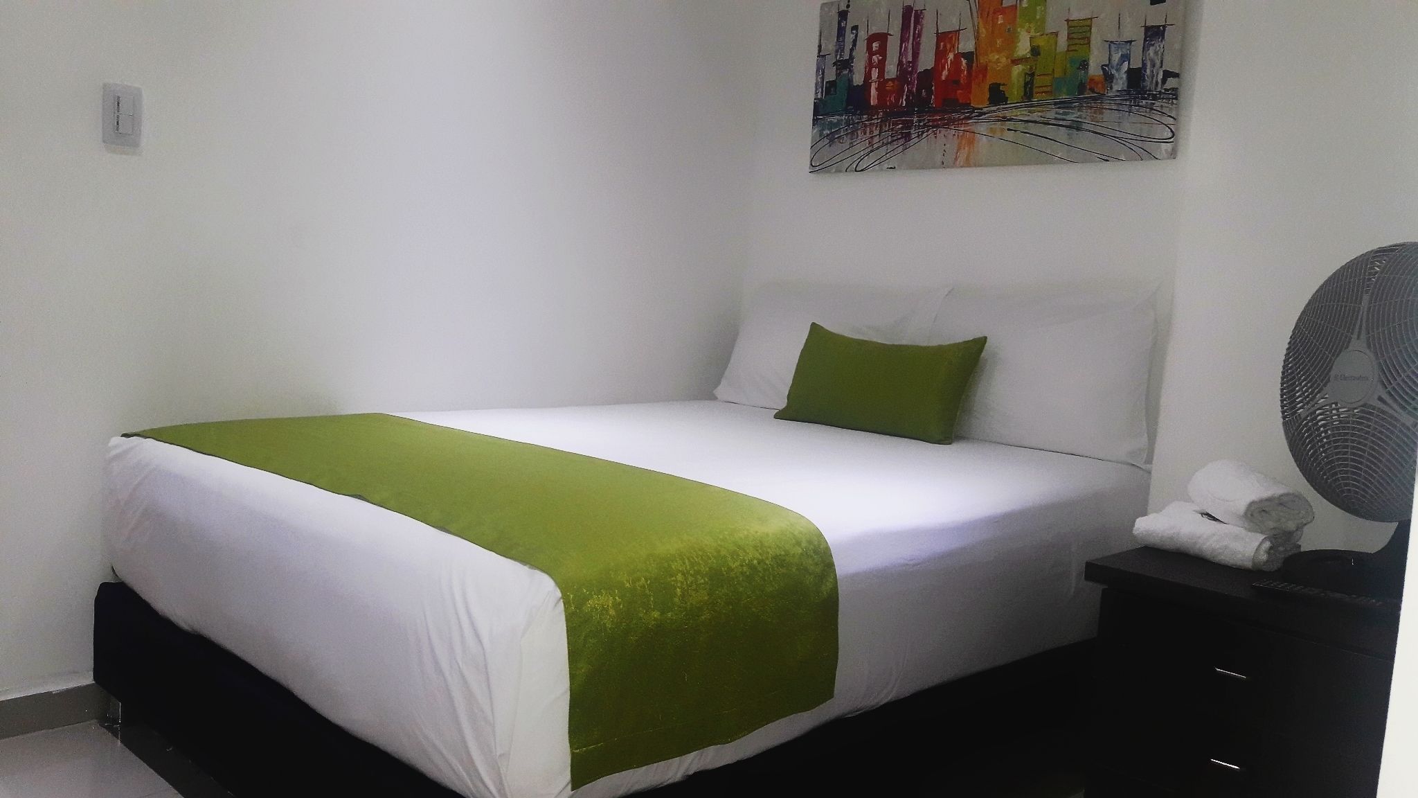 A neatly made bed with white and green bedding, a green pillow, and a black nightstand with a white towel and a fan.