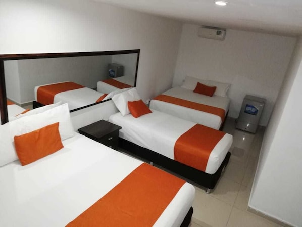 Habitación de hotel con tres camas, cada una con ropa de cama blanca y detalles naranja. Hay un espejo grande en una pared y un cesto de basura en la esquina.