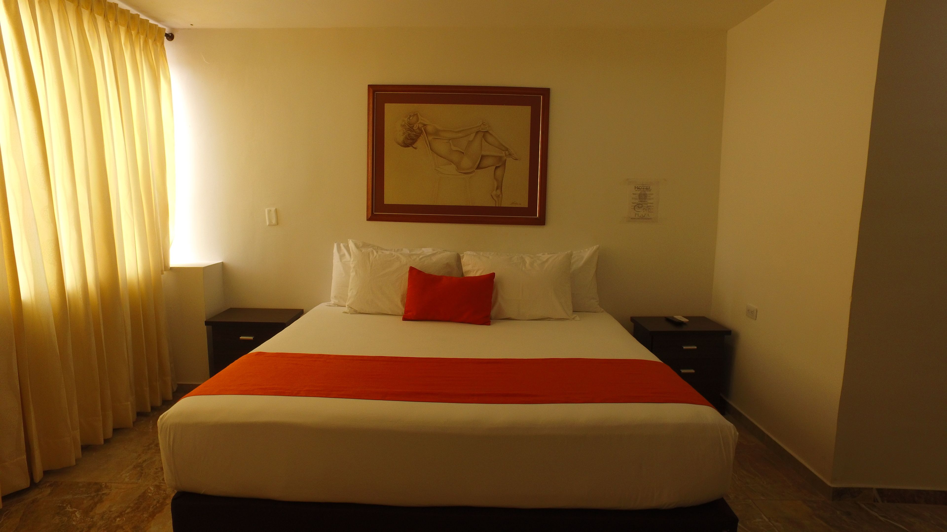 A hotel room with a large bed, a nightstand, a framed artwork above the bed, and a window with curtains.