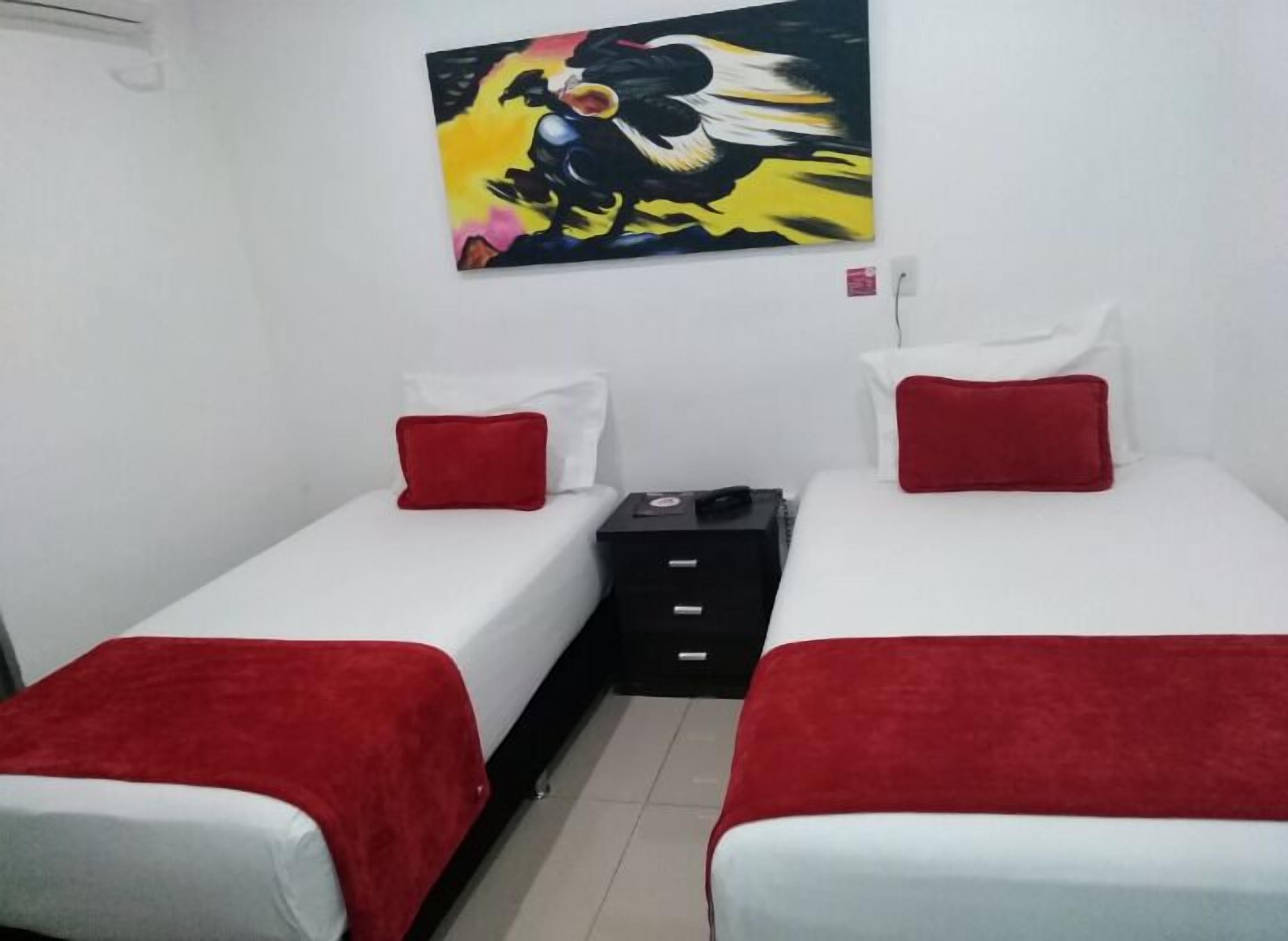 A double room with two beds, each with a red pillow and white bedding. There is a painting on the wall above the beds.