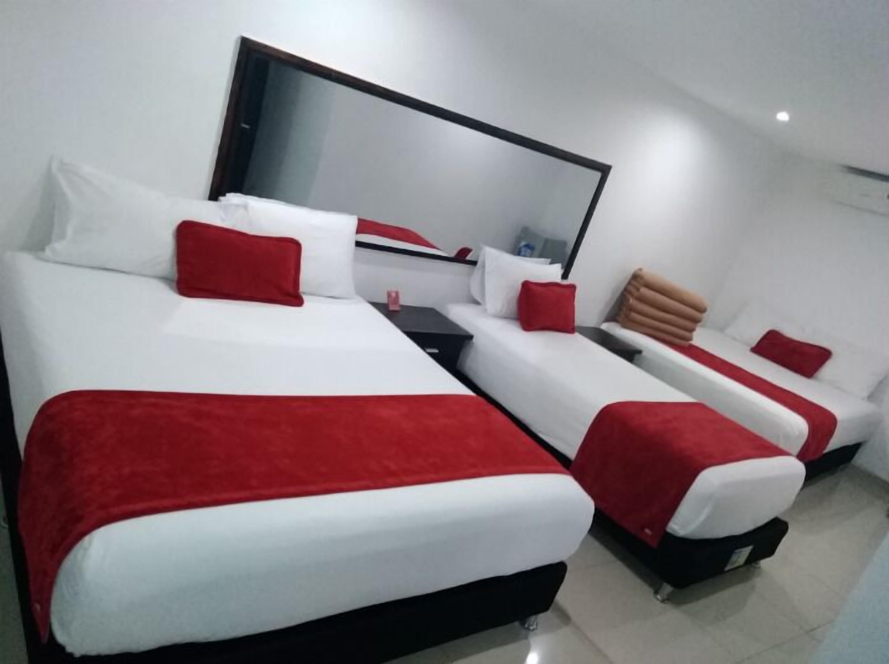 Habitación de hotel con tres camas, cada una con ropa blanca y detalles rojos. Hay un espejo grande sobre las camas y un televisor de pantalla plana sobre un pedestal.