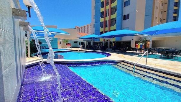 A swimming pool with a water feature, surrounded by outdoor seating and a multi-story building in the background. - Thermas das Caldas - Achei Férias (Caldas Novas)