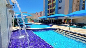 Una piscina con elemento d'acqua, circondata da posti a sedere all'aperto e un edificio a più piani sullo sfondo.