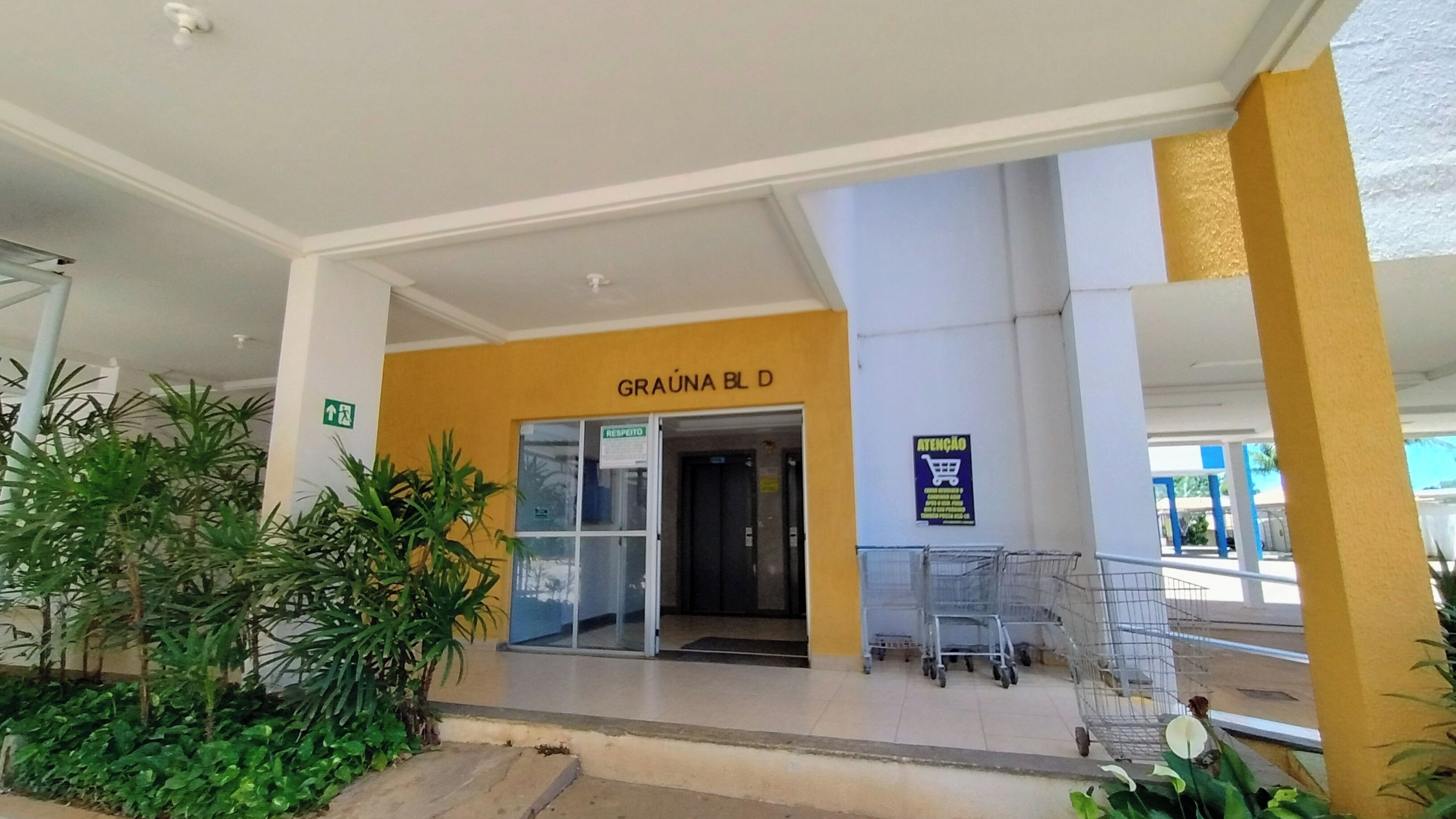 Entrada de um edifício com esquema de cores amarelo e branco, uma placa com a inscrição "GRAÚNA BL D" e um carrinho de compras próximo à entrada.