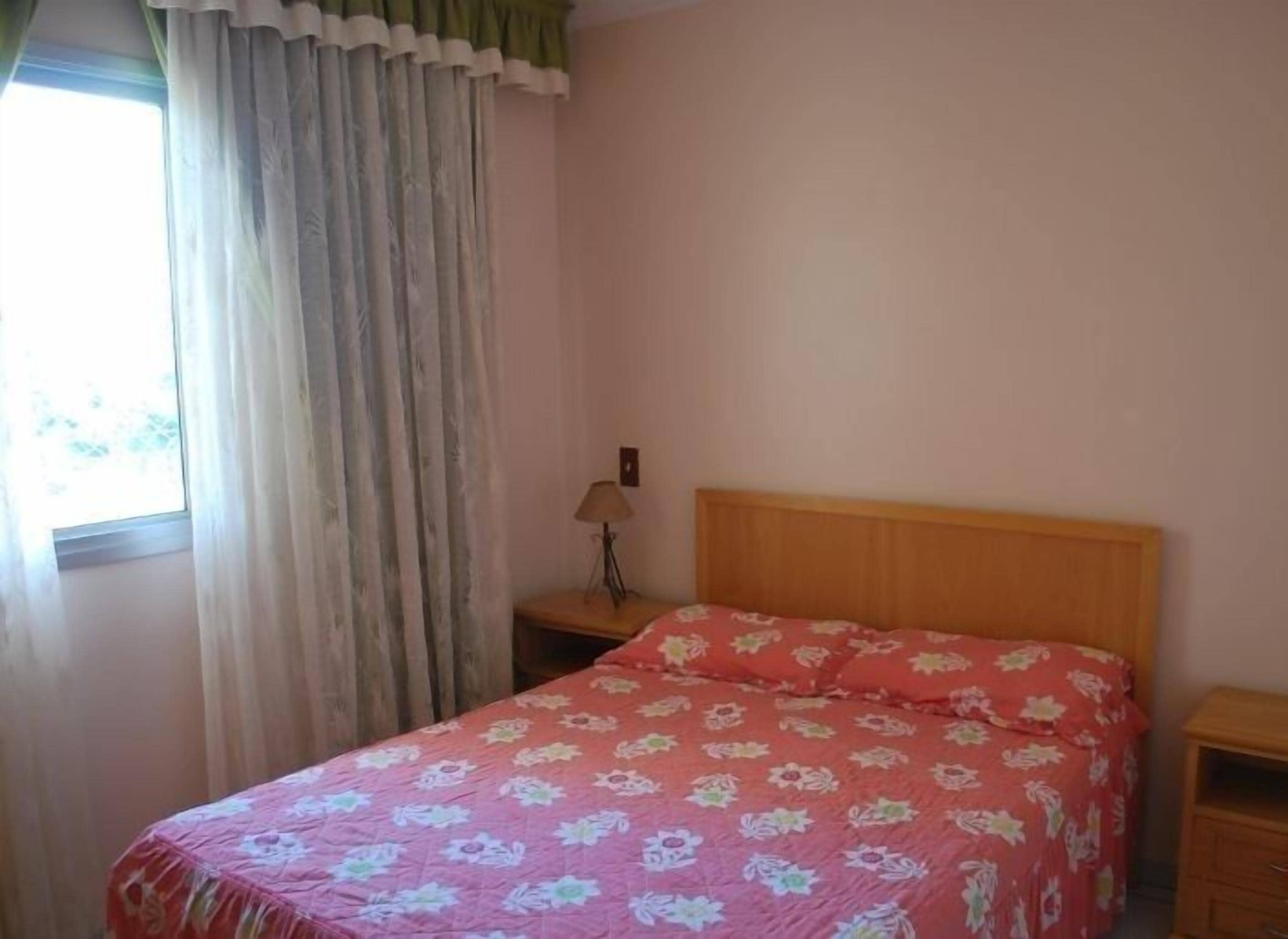 Quarto com cabeceira de madeira, colcha floral, luminária de cabeceira e janela com cortinas.