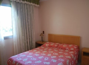 A bedroom with a wooden headboard, a floral bedspread, a bedside lamp, and a window with curtains.