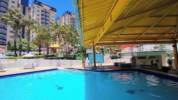 Piscina com área coberta, cercada por palmeiras e um edifício com vários andares.