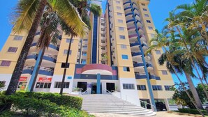 Un hotel de varios pisos con una entrada central, rodeado de palmeras y un cielo azul despejado.